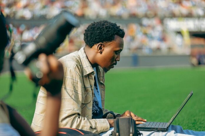 Jornalista negro trabalhando em seu notebook. Ao fundo, é possível ver as arquibancadas e o gramado de um estádio de futebol.